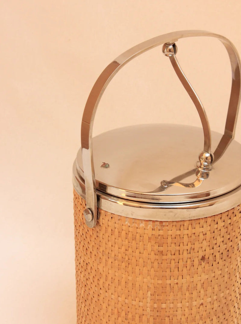 The Ratan Ice Bucket from Boga Avante Shop features a cylindrical wicker exterior with a shiny metal lid and an elegantly curved handle, set against a beige background. Its blend of functionality and minimalist design makes it an eye-catching sculptural piece.