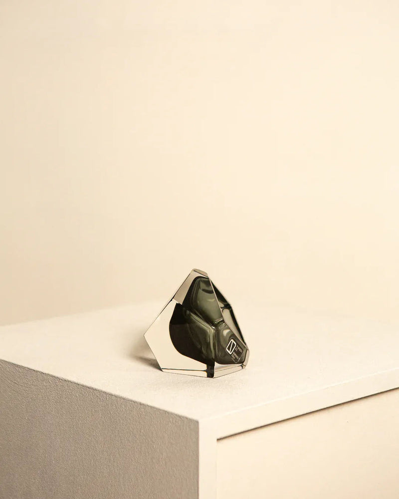 A small, geometrically-shaped black Treaptyque handbag with a metallic frame is placed on a white textured pedestal against a neutral beige background. Reminiscent of Flavio Poli’s designs in Murano glass, this "Diamant" Vide Poche by Flavio Poli for Seguso 60's features a sleek, angular design with a single handle and minimalistic aesthetic.