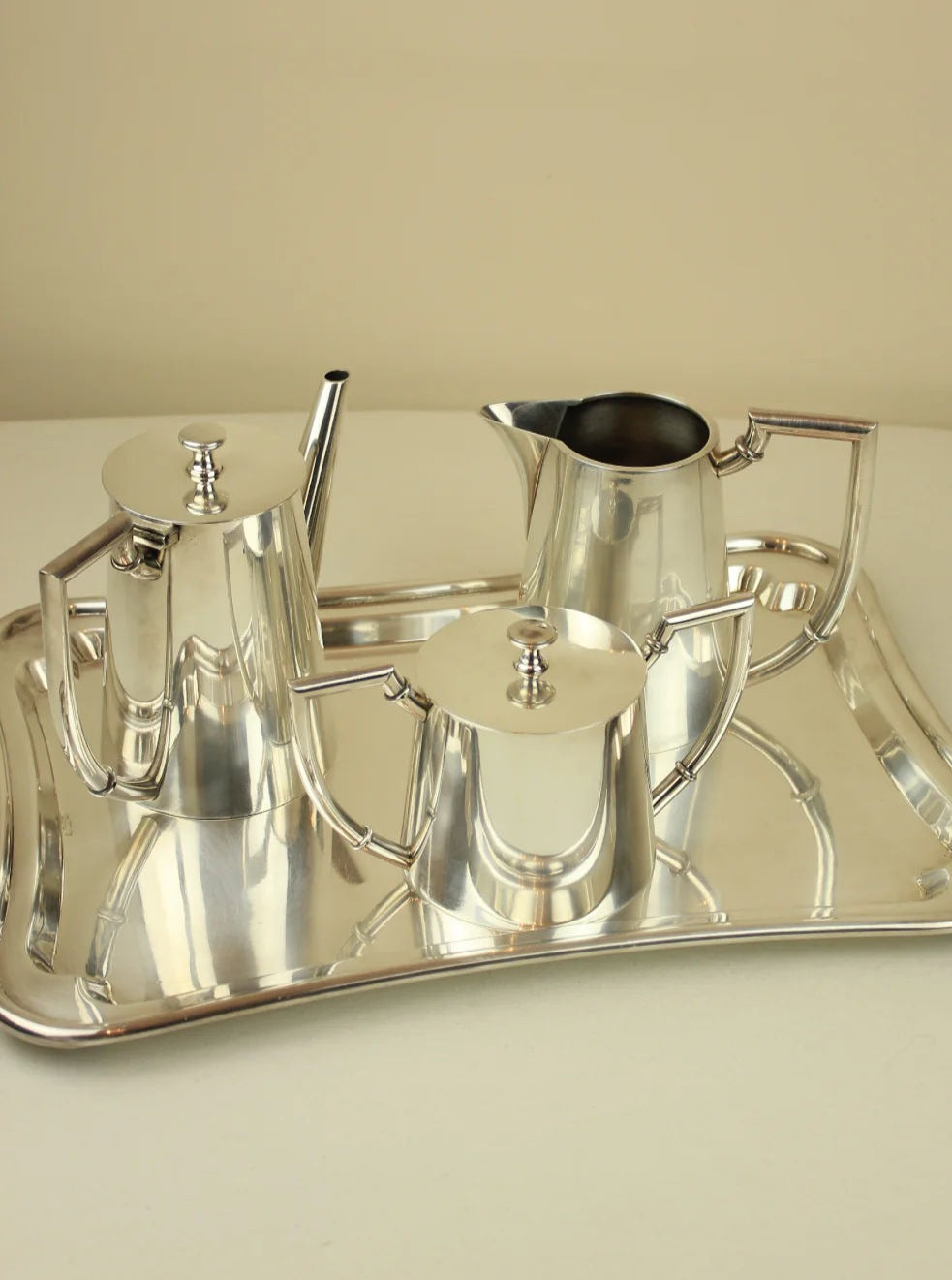 A polished silver Boga Avante Shop Art Deco Tea/Coffee Service on a tray, including a teapot, sugar bowl, and creamer, all with lids and classic designs, displayed on a beige background.