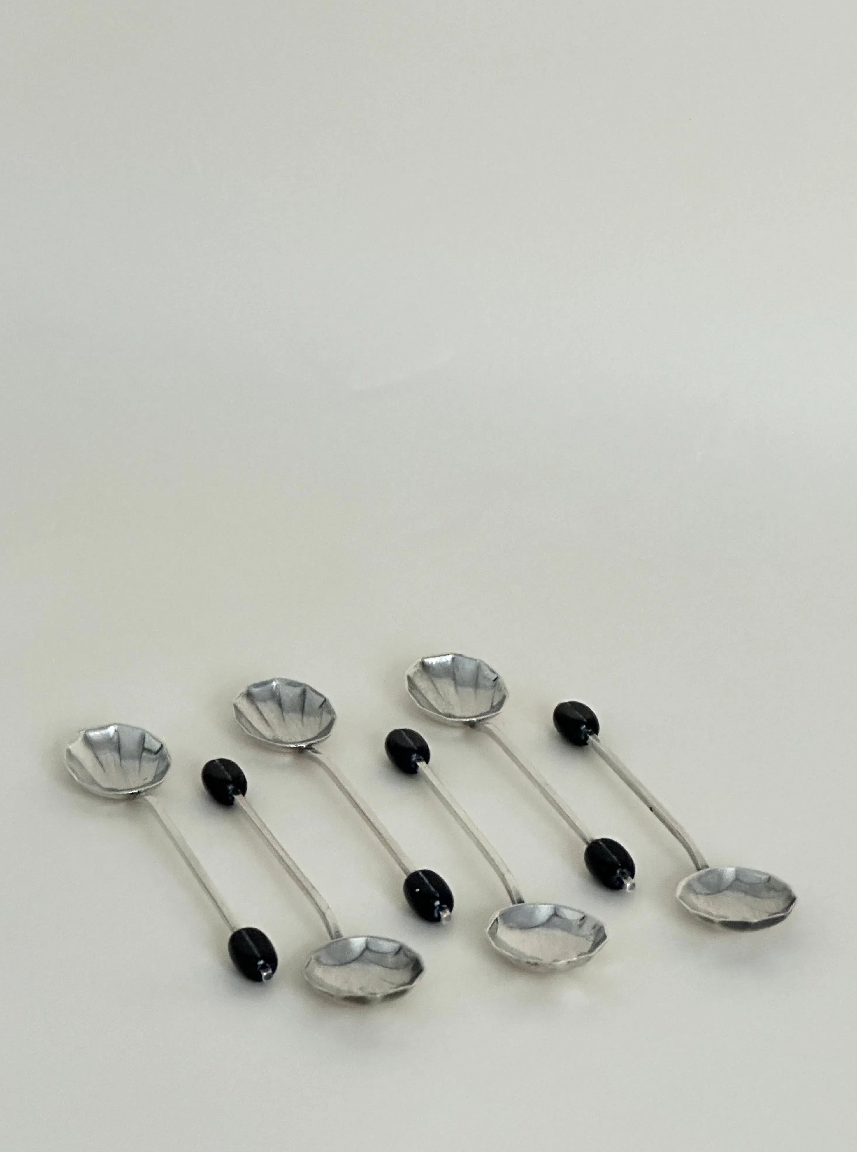 A collection of eight silver plate cocktail picks with decorative ends, featuring alternating designs of black ovals and transparent faceted crystals, arranged diagonally on a light background by Aesthete Label.