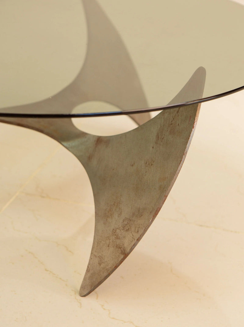 A close-up of the Propeller Circular Coffee Table by Knut Hesterberg from Treaptyque showcases its modern aesthetic, with a glass top and an artistic triangular wooden base resting on a light-colored floor. The sleek lines and smooth finish contribute to its stylish and minimalist appearance.