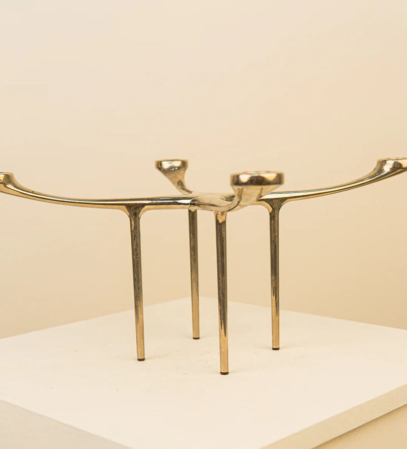 A golden abstract sculpture featuring two elongated, arched forms with a sleek finish, resembling stylized figures or antelope horns, displayed on a white pedestal against a neutral background inspired by Scandinavian design by Treaptyque.