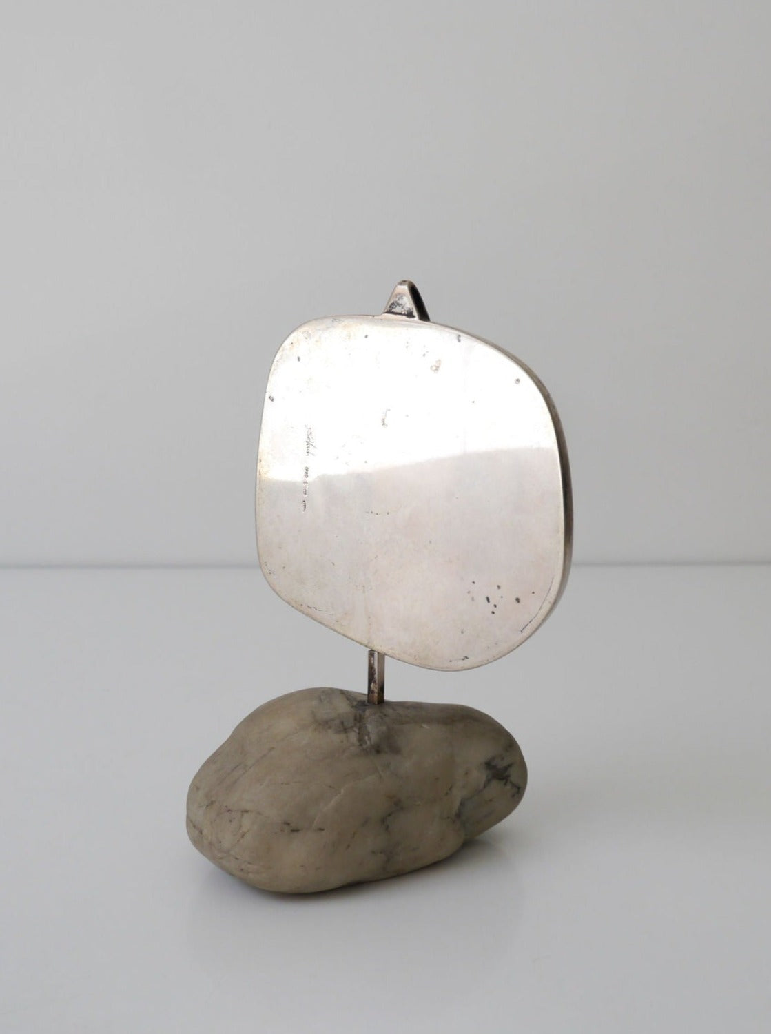 A minimalist sculpture featuring a polished De Vecchi Milano silver square attached to a stem, which is anchored in a smooth, irregularly shaped stone base on a plain white background by Collection apart.