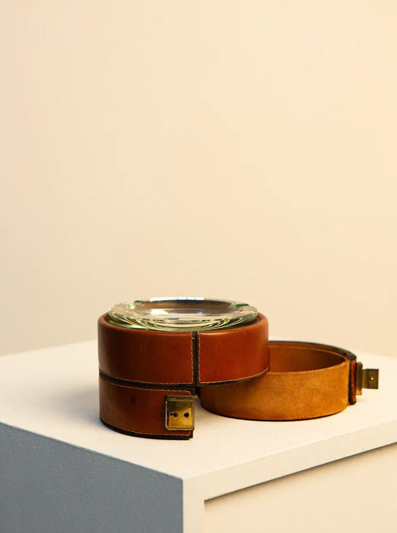 A Treaptyque Italian Ashtray in Brown Leather and 60's Crystal rests on a white pedestal against a plain, light-colored background. The sturdy and well-crafted brown leather holder, featuring brass clasps, showcases the timeless craftsmanship of the 1960s, adding an elegant aesthetic to the ashtray.