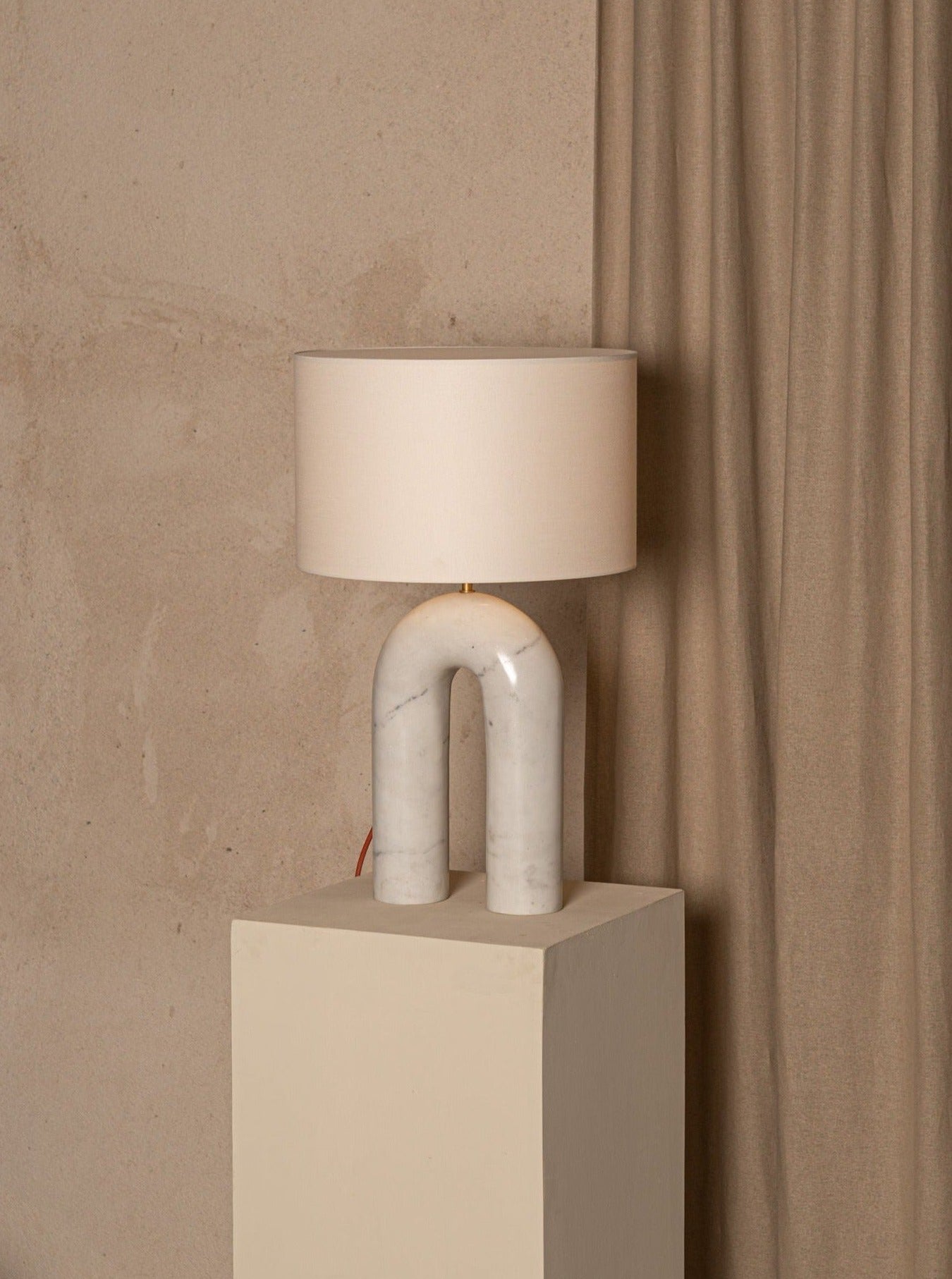 A unique Arko White Marble Lamp by Simone & Marcel, with a marble base shaped like an arch, supporting a cylindrical cotton lampshade, placed on a beige cube pedestal against a textured beige wall.