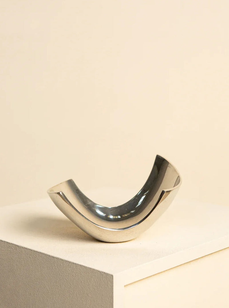 A modern, metallic crescent-shaped 80's "U" Italian Fruit Basket from Treaptyque, resembling a silver metal basket, sits on a light-colored pedestal against a soft beige background, displaying a sleek, minimalist design.