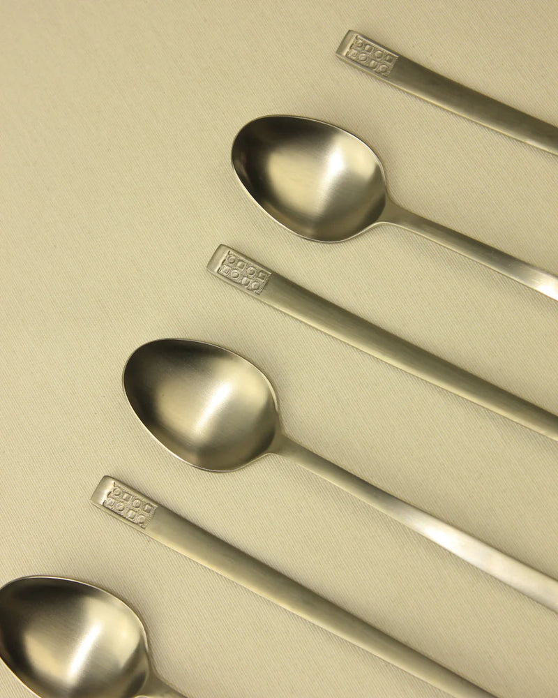 Four Boga Avante Shop 70s Style Spoons with decorative handles lined up diagonally on a beige textured background.