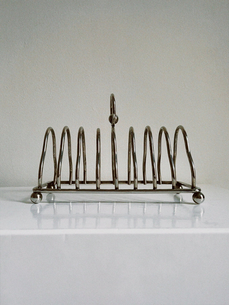 A sleek design Manushi Art Deco Toast Rack with six vertical holders and a central looped handle, placed on a light-colored surface against a neutral background.
