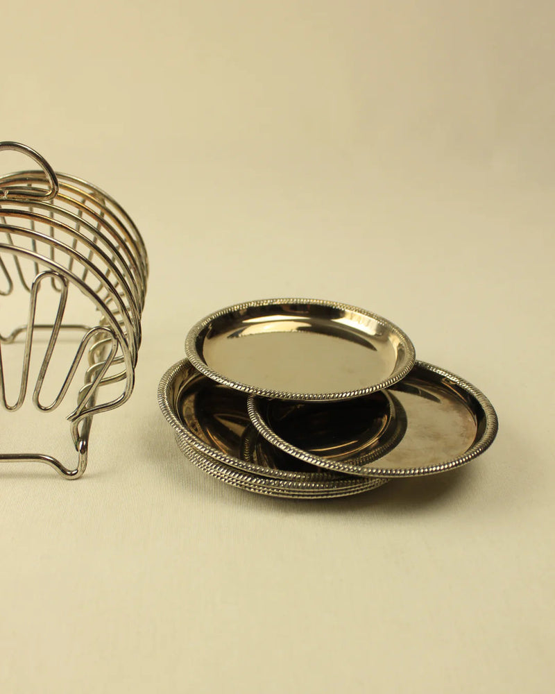 Three shiny, silver Boga Avante Shop bread plates with intricate beaded edges next to a silver Art Nouveau style wire basket, all placed on a beige surface.