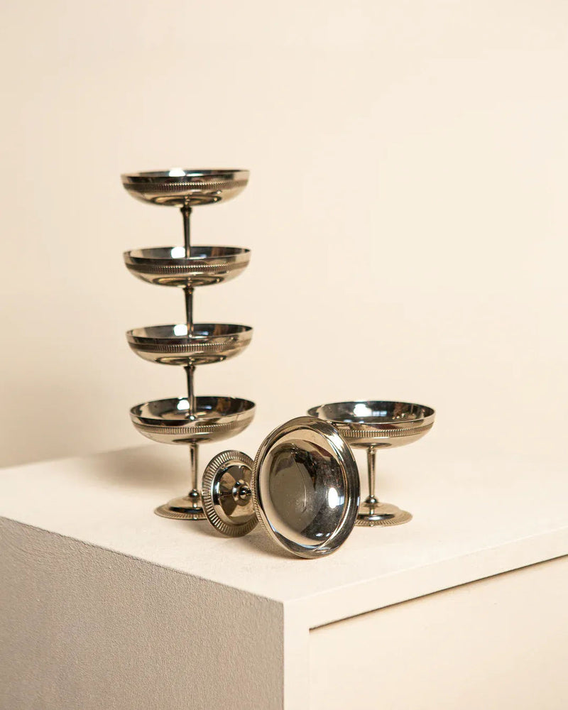 A set of elegant Treaptyque silver candle holders displayed on a white pedestal against a beige background embodies vintage glamour. The tallest holder features a stack of five dishes, while others are simpler in design.