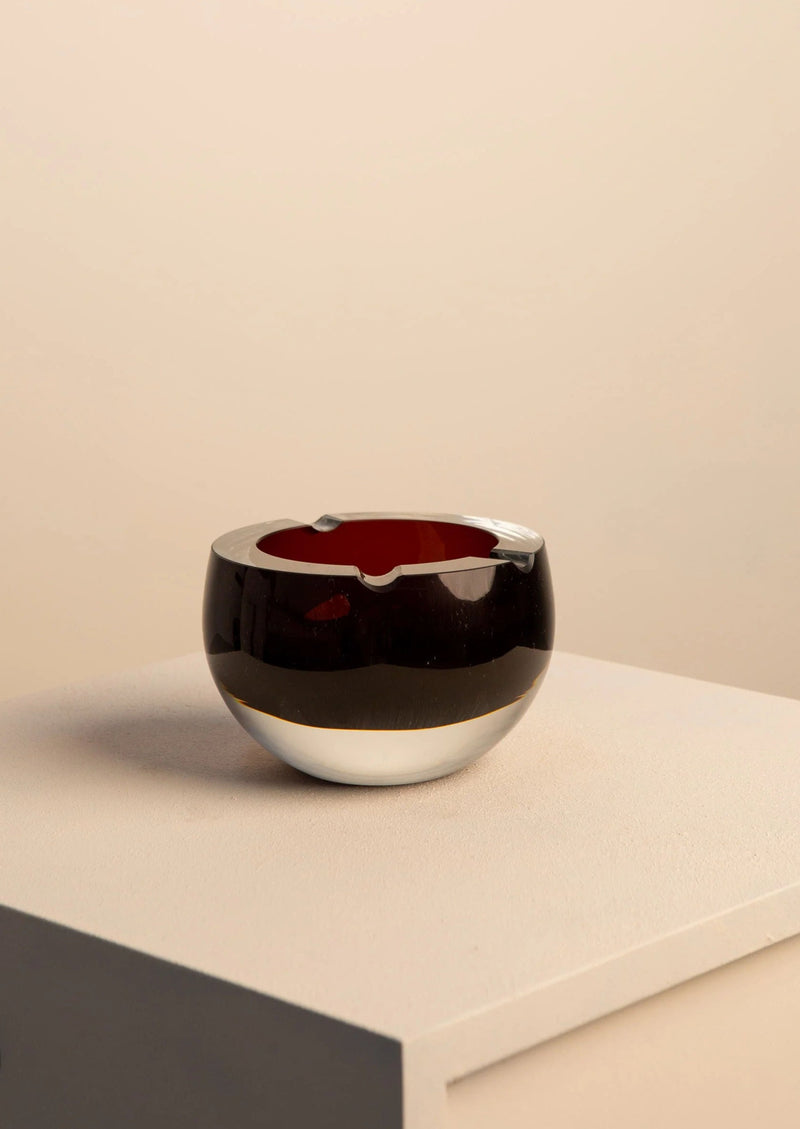 A large, round Murano glass ashtray by Treaptyque, featuring a dark exterior and red interior, rests on a white pedestal. The backdrop is a soft beige, creating an elegant and minimalist setting.