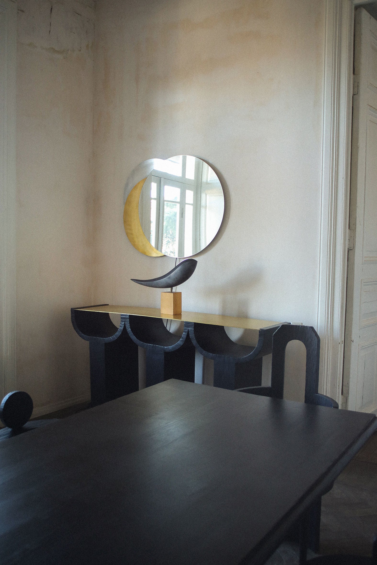 A minimalist room with a sleek brass table, unique sculptural black chair, and an Eclipse Mirror from Rooms Studio against a wall near a window, casting soft natural light.