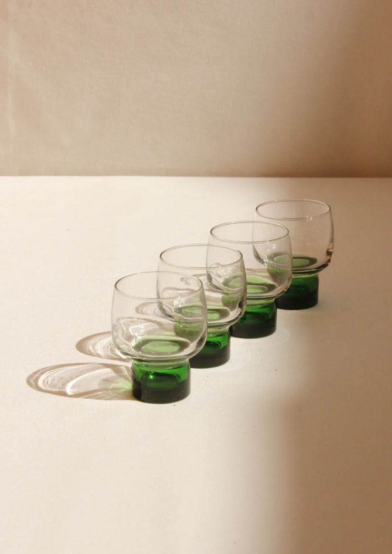 Four Boga Avante Shop clear glass French liqueur glasses with green bases and curved handles, lined up diagonally on a beige surface against a plain, light-colored background.