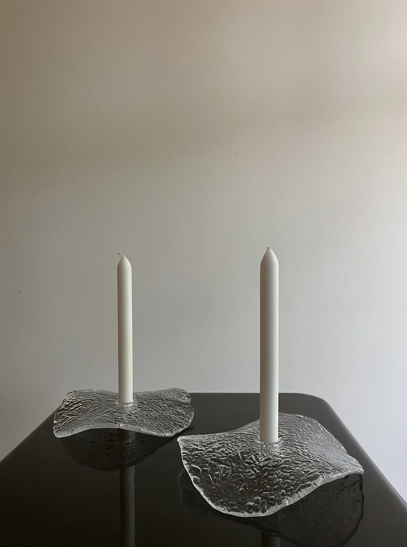 Two white candles stand upright in the Uno Westerberg Candle Holder Set by Les Objets Amsterdam atop a reflective black surface. The background is minimalistic with a light grey wall, emphasizing the serene and elegant arrangement.