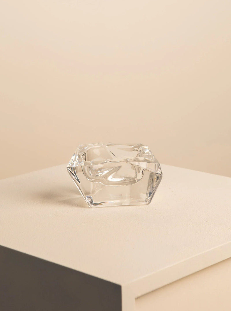 A small, clear glass vase with an abstract, geometric design is placed on a simple, off-white platform against a beige background. The vase's polished, reflective surface brings to mind the elegance of Treaptyque's Free-Form Crystal Ashtray by Daum from the 60s and appears to be empty.
