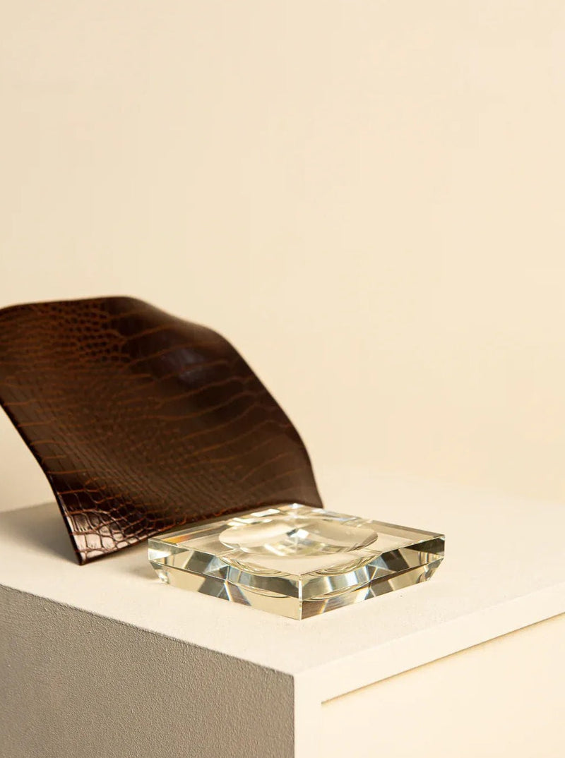 An Italian crystal Treaptyque ashtray on a white plinth, next to a folded dark brown textured Treaptyque item, set against a neutral beige background. The ashtray catches the light, creating a sparkle effect.