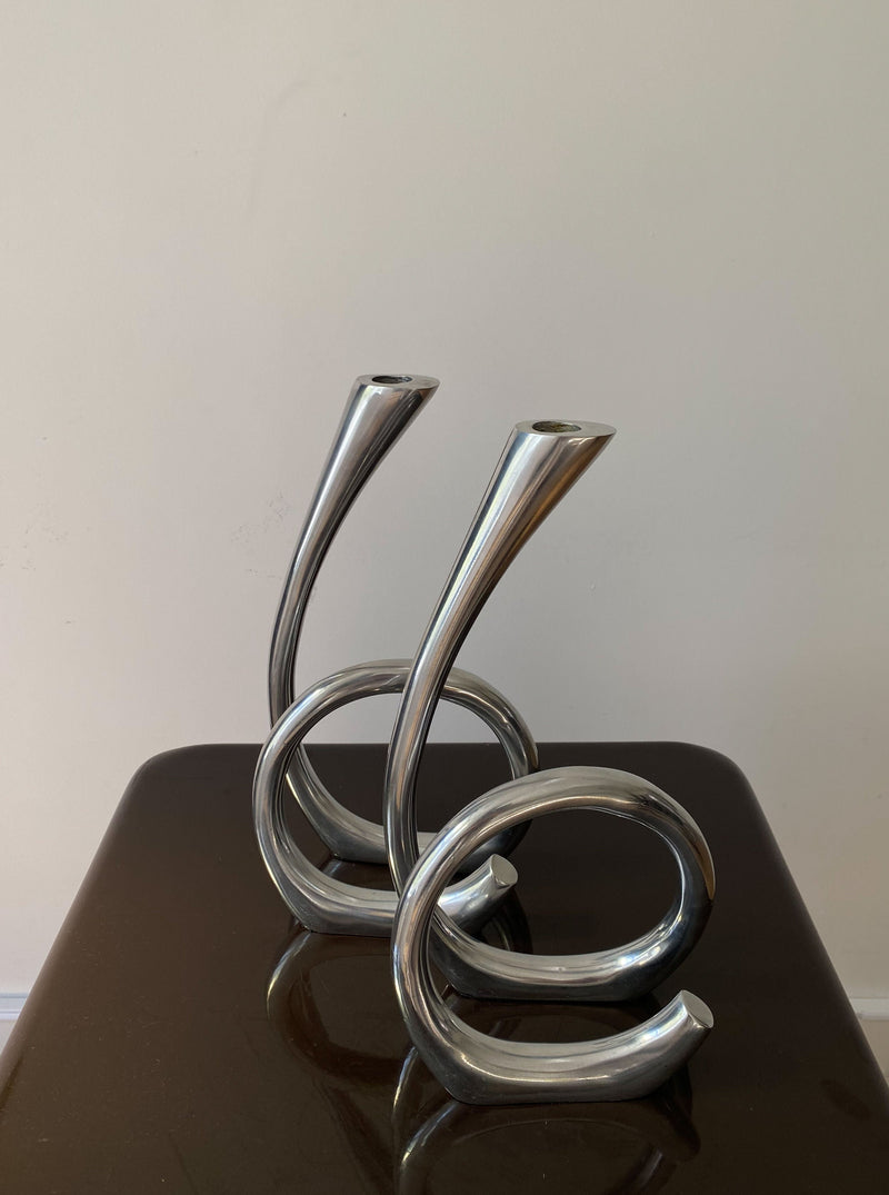 Two Vintage Curly RVS Candle Holders with twisted metal designs rest on a glossy, dark surface against a pale wall by Les Objets Amsterdam.