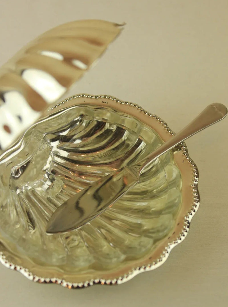 The Boga Avante Shop's Shell Shaped Butter Dish features a polished silver finish with a matching lid, slightly ajar to reveal its inner compartment. A sleek, decorative butter knife with a smooth handle rests inside the dish, positioned diagonally. The dish boasts a reflective, shiny surface that beautifully complements the intricately designed knife.