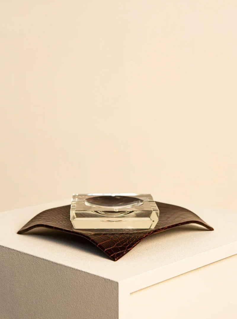 A Treaptyque crystal ashtray in Crystal and Brown Italian Crocodile Leather 80's rests on a textured brown leather mat, which is placed on a beige box against a plain off-white background, creating a minimalist aesthetic with an Italian crystal detail.