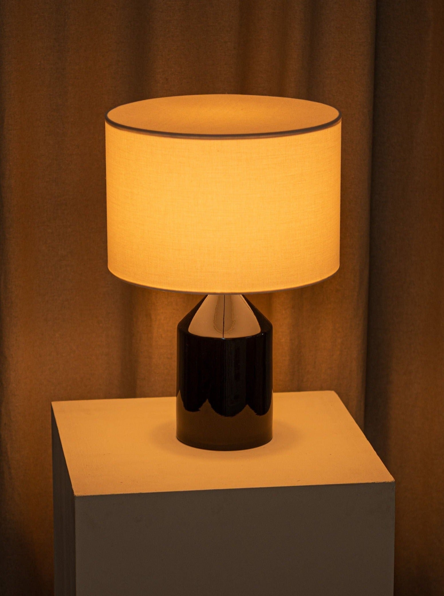 A warmly lit Simone & Marcel Josef Black Ceramic table lamp with a cotton drum lampshade, casting a soft glow on a draped beige curtain background. The lamp's base is glossy, dark, and partially reflective, standing on a