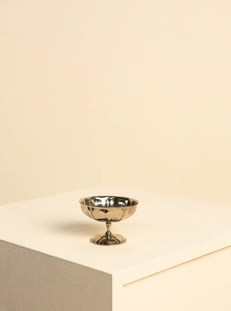 A small, shiny, silver **Set of 8 - 70's Stainless Steel Ice Cream Bowls** from **Treaptyque** sit on a beige podium against a light beige background. Reminiscent of retro design and French artisanal craftsmanship, the ice cream bowls feature a wide, shallow bowl and a narrow stem with a flat base, reflecting light and casting soft shadows.