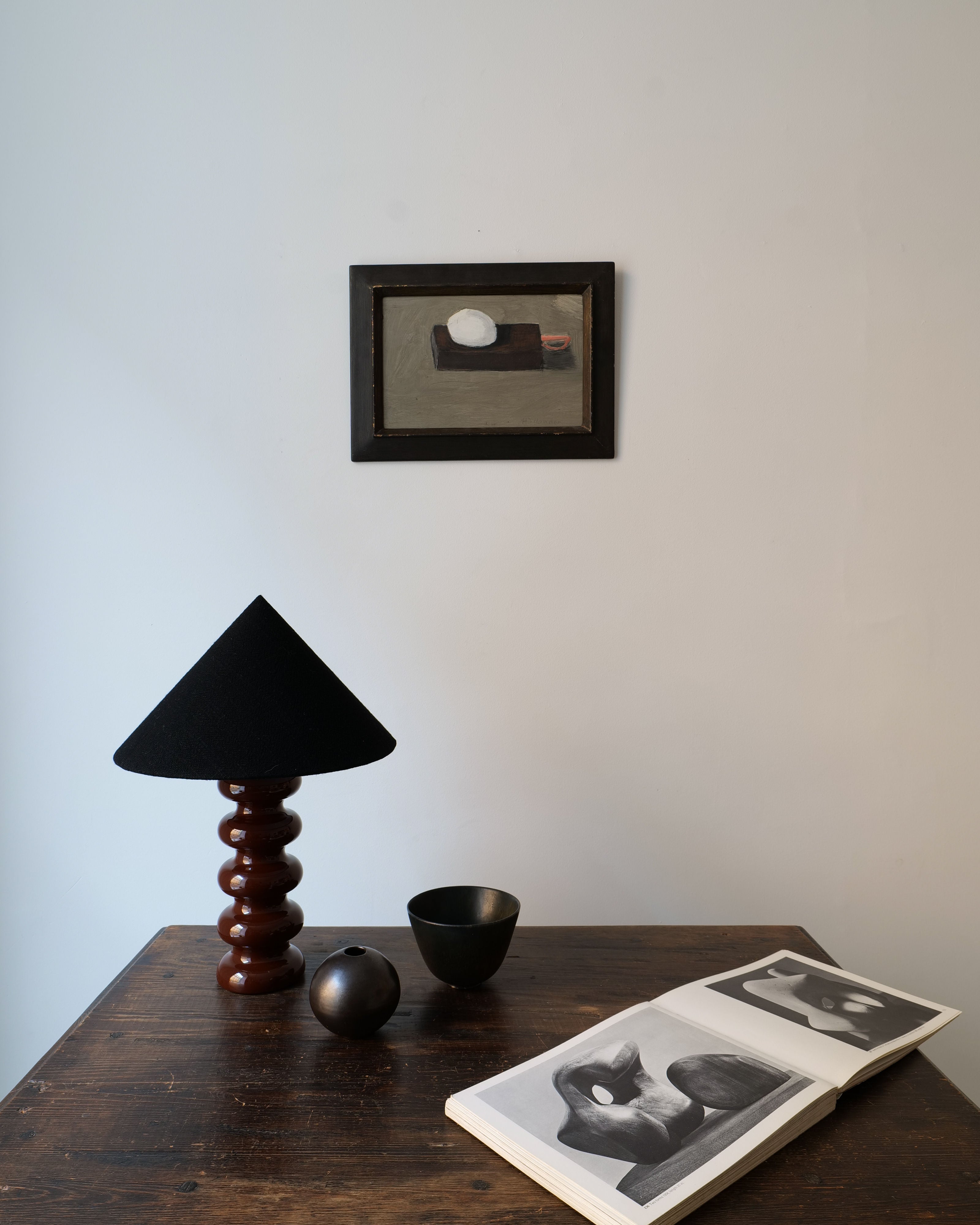 A minimalist interior setup with a wooden table displaying a Collection apart Sculptural Ceramic Lamp, two dark spherical objects, and an open book with black and white photographs. A framed painting hangs on the pale wall above.