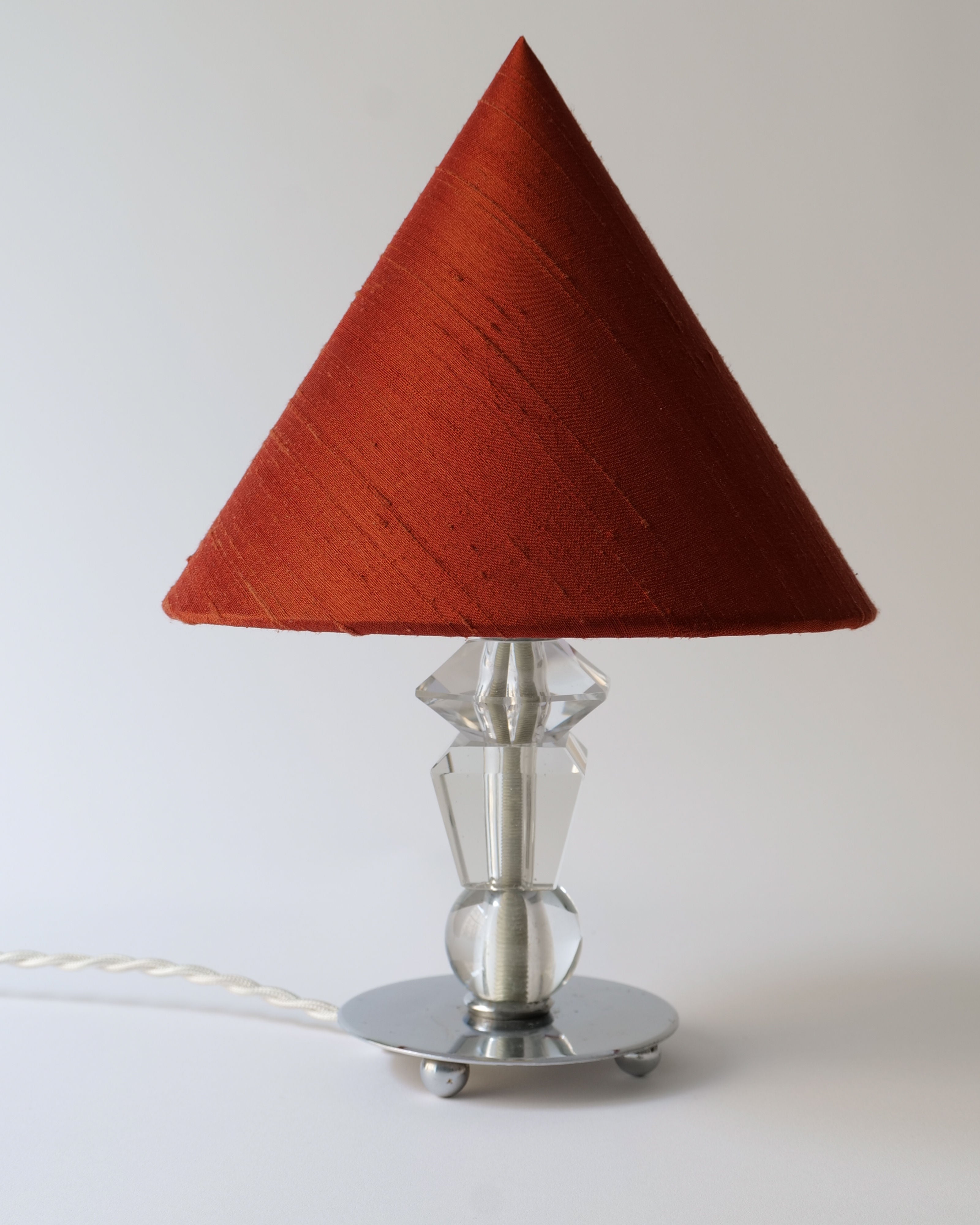 A contemporary boudoir bedside lamp with a striking red conical shade, mounted on a transparent glass base with a metallic stem and circular platform, accompanied by a white woven cable: The Deco Crystal Table Lamp by Collection apart.
