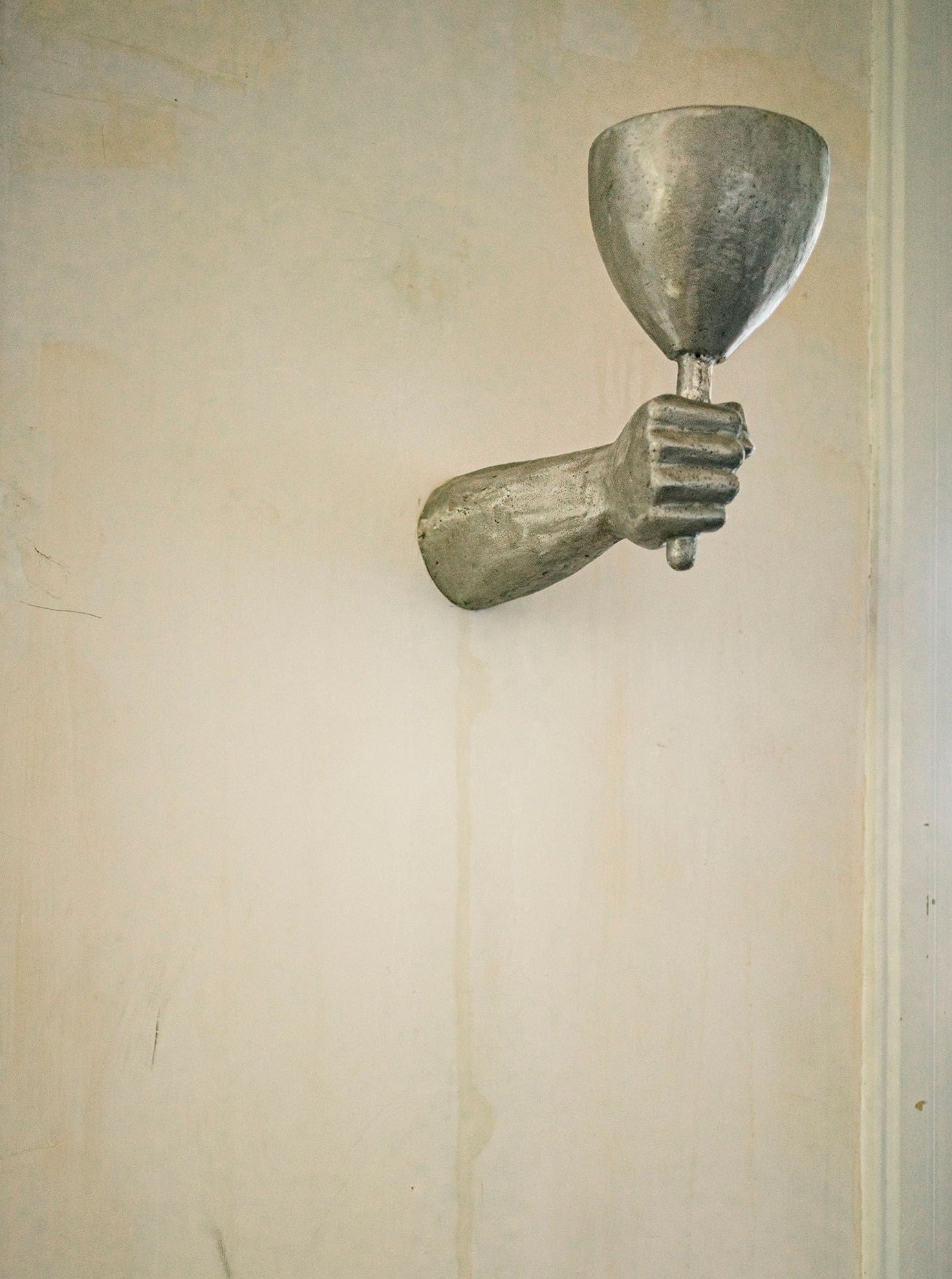 A sculpture of a human hand protruding from a wall, holding a wide-brimmed chalice, set against a plain, lightly textured backdrop with a vertical crack. This piece is crafted from cast Dracula Wall Scone Aluminium by Rooms Studio.