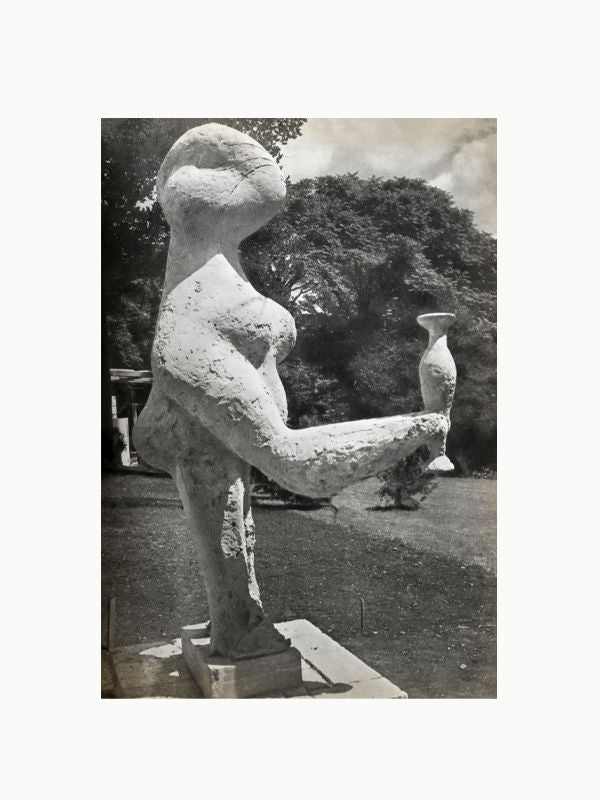 A vintage black-and-white photo of an abstract Maison Plage sculpture from "The Sculptures of Picasso (1st Edition, 1949)" that features a humanoid figure with smooth, flowing lines, holding a vase or jug in its raised arms, set outdoors under a tree.