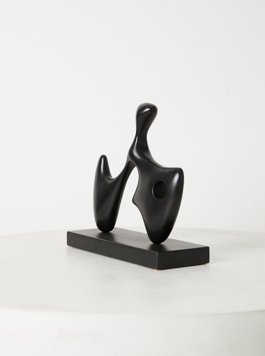 A black, abstract wood sculpture resembling a stylized figure sits on a rectangular base, displayed against a simple, light background.