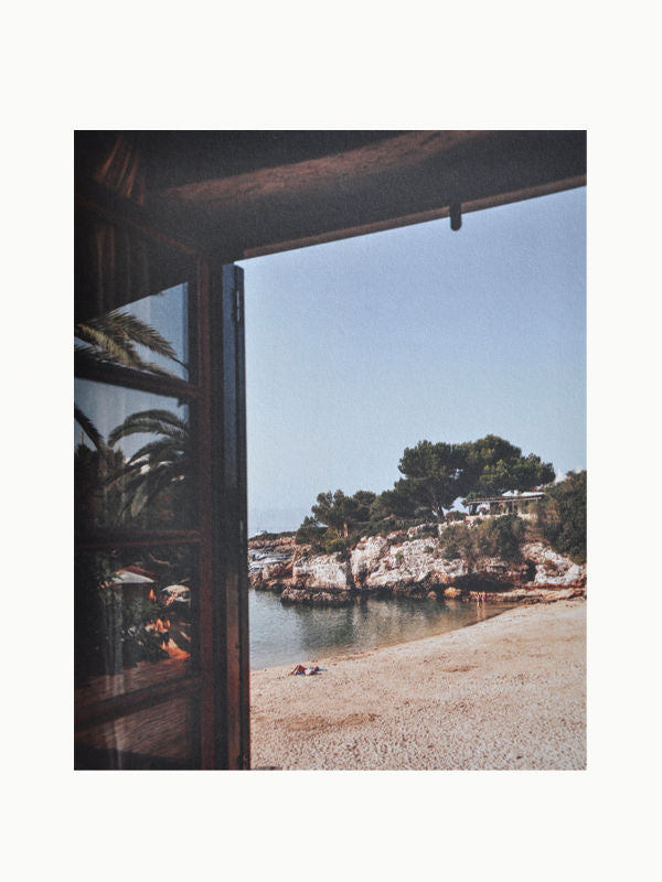 View from an open doorway showing a serene beach with light-colored sand, rocky formations, and a calm sea. Trees and greenery adorn the rocky cliffs in the background under a clear blue sky. Nearby, The Orange Set of 3 Interior Design and Art Books by Maison Plage is thoughtfully arranged, adding an element of creative inspiration to the scene.