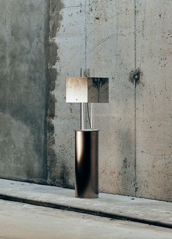The Brajak Vitberg Metal Floating Table Lamp, embodying contemporary design, features a cylindrical base and mirrored square element that artfully reflects its surroundings. The smooth concrete floor enhances the lamp's minimalist aesthetic as it stands in front of a textured concrete wall.