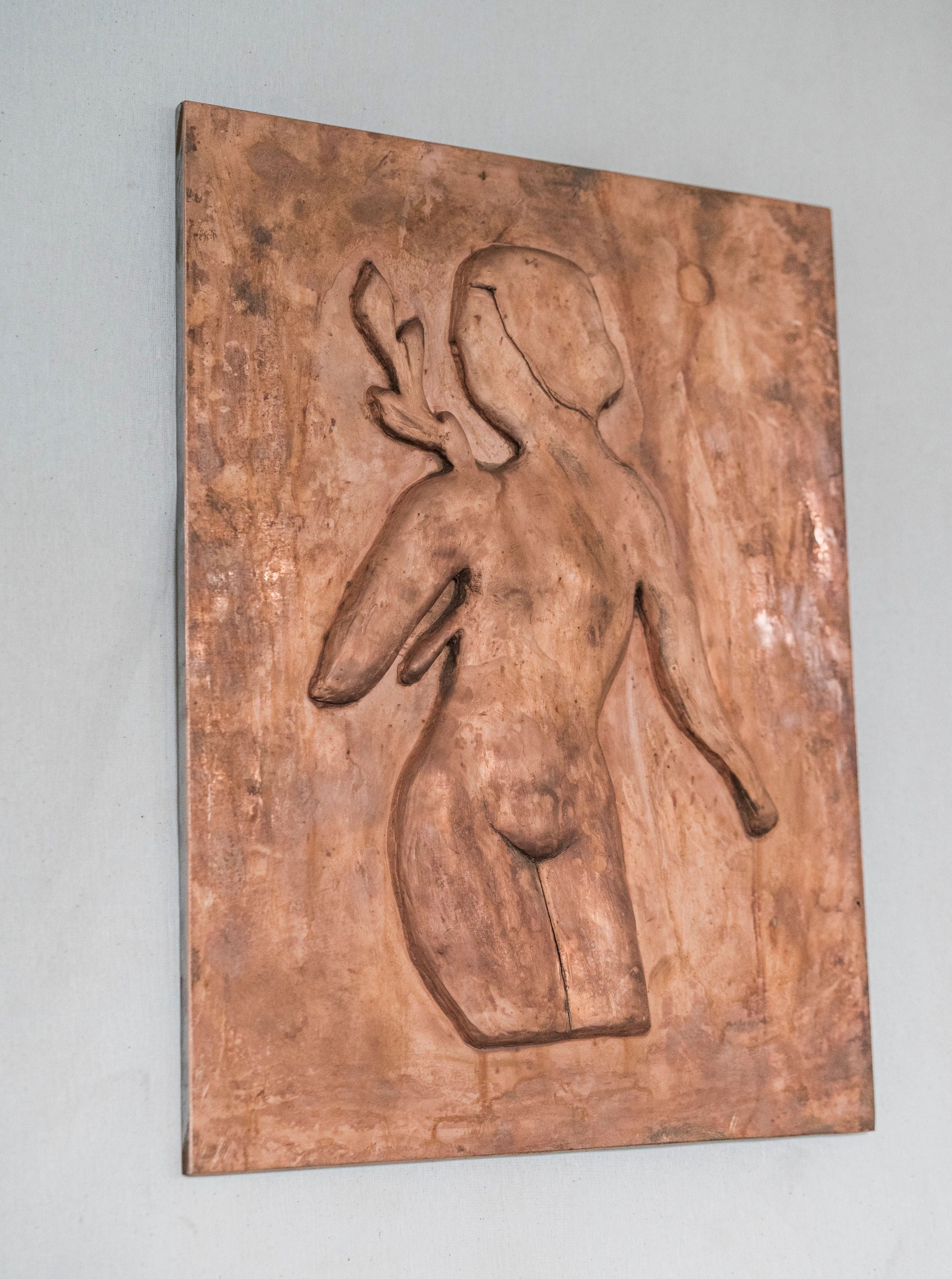 An embossed Woman with Laurel leaves Chikanka wall sculpture of an abstract, stylized human figure on a copper-toned plaque mounted on a white wall by Rooms Studio.