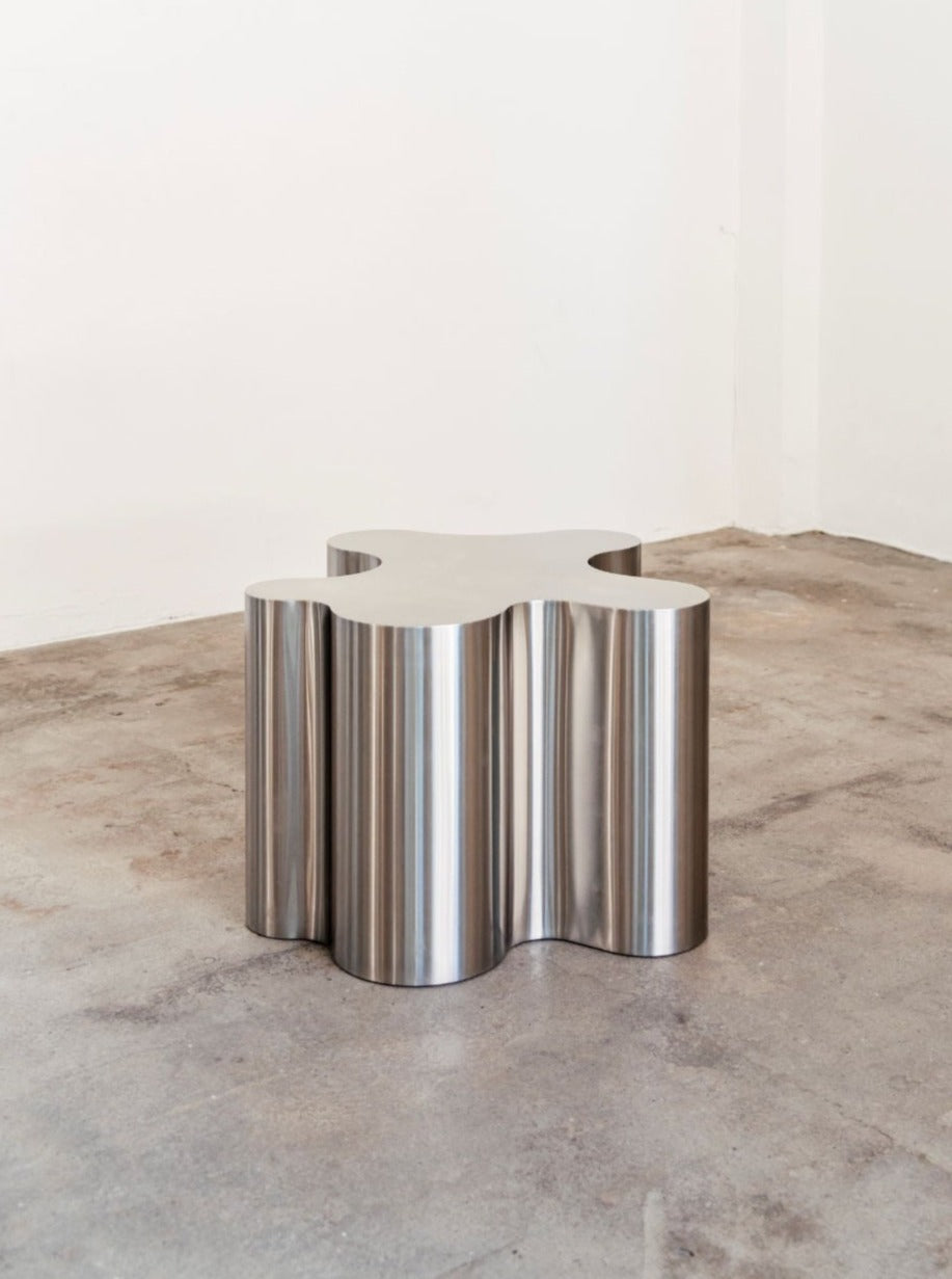A modern, metallic Silver Root - Low stool with a sculptural steel base sits on a concrete floor against a plain white wall. The stool's surface is polished, reflecting subtle light. (Caia Leifsdotter)