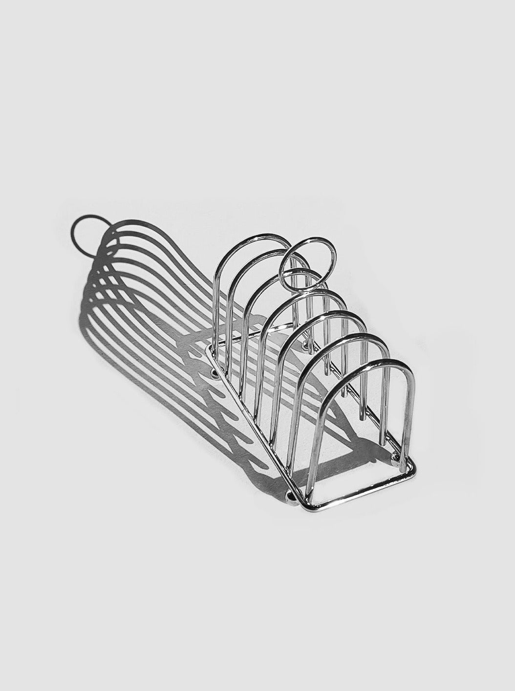 A modern stainless steel Art Deco Toast Rack from Les Objoies with four slots, viewed on a white background, casting geometric shadows.