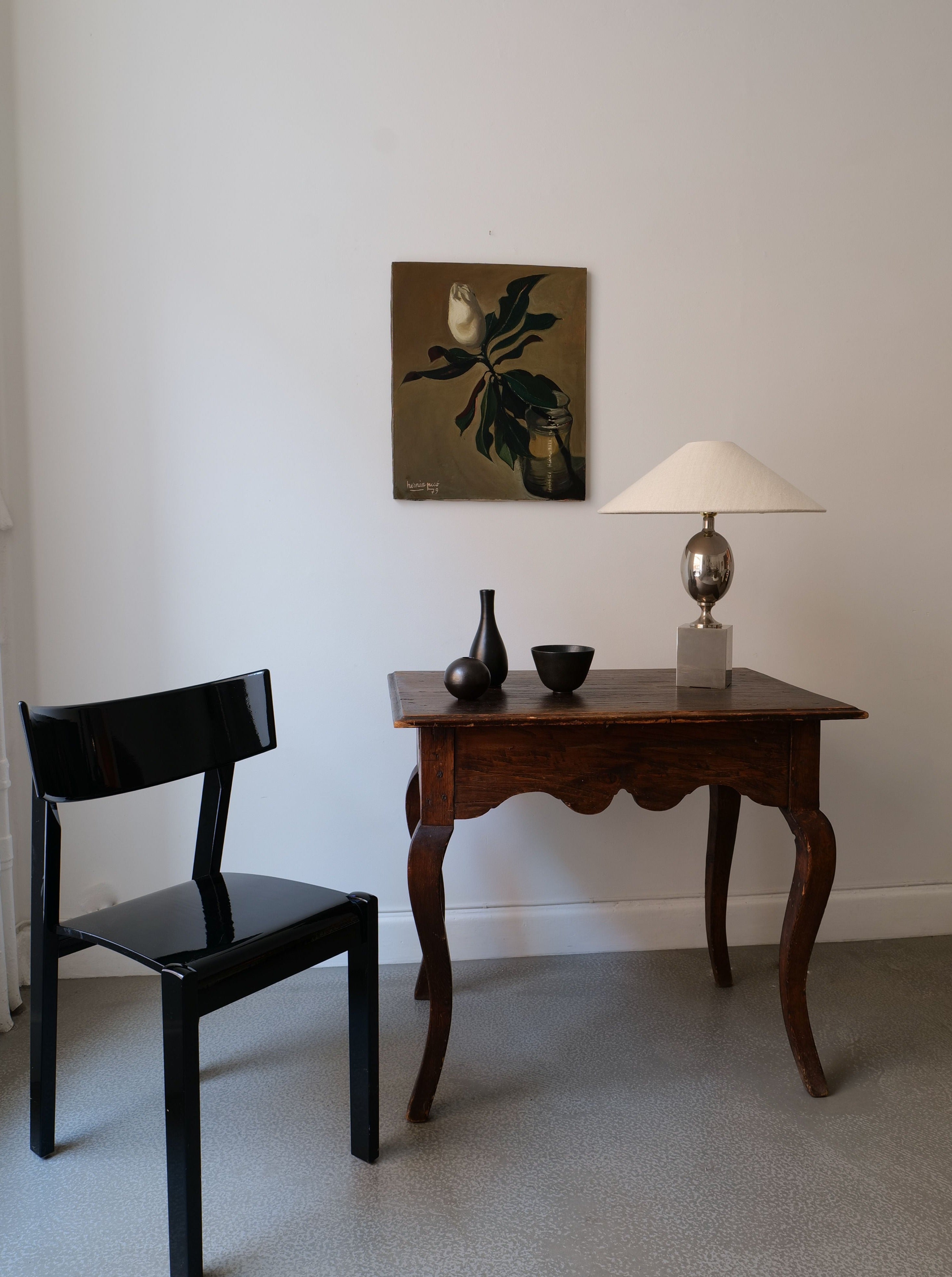 A simple interior setting featuring a dark wooden chair beside an antique wooden table with a sleek table lamp and two decorative vases. There is a Collection apart Magnolia Grandiflora 1979 on canvas floral painting on the wall above the table.