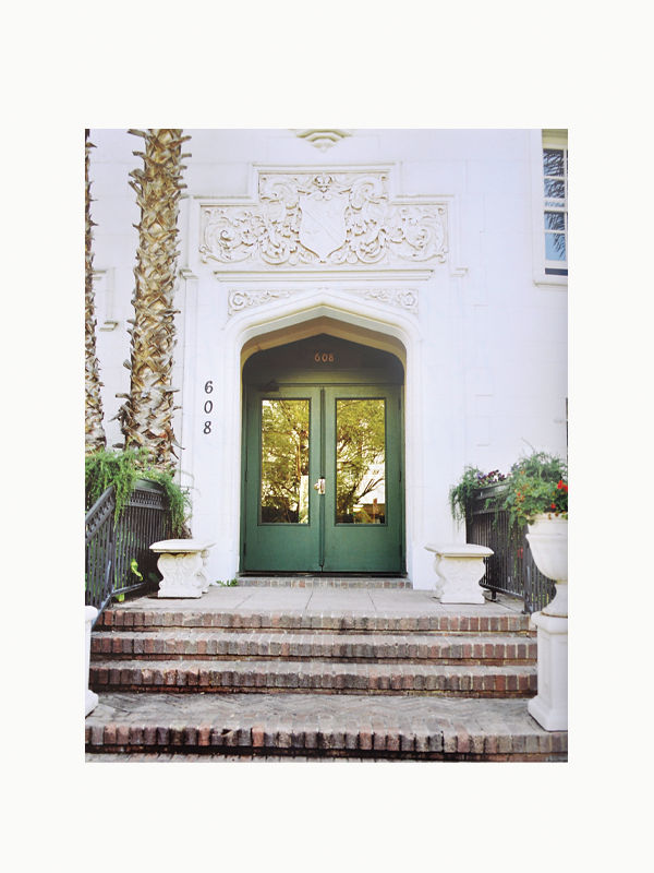 An elegant doorway in Art Deco style, with ornate white carvings above, reflecting glass doors at number 608, flanked by planter pots and a palm tree, ascedned by Preservation of Style from Maison Plage.