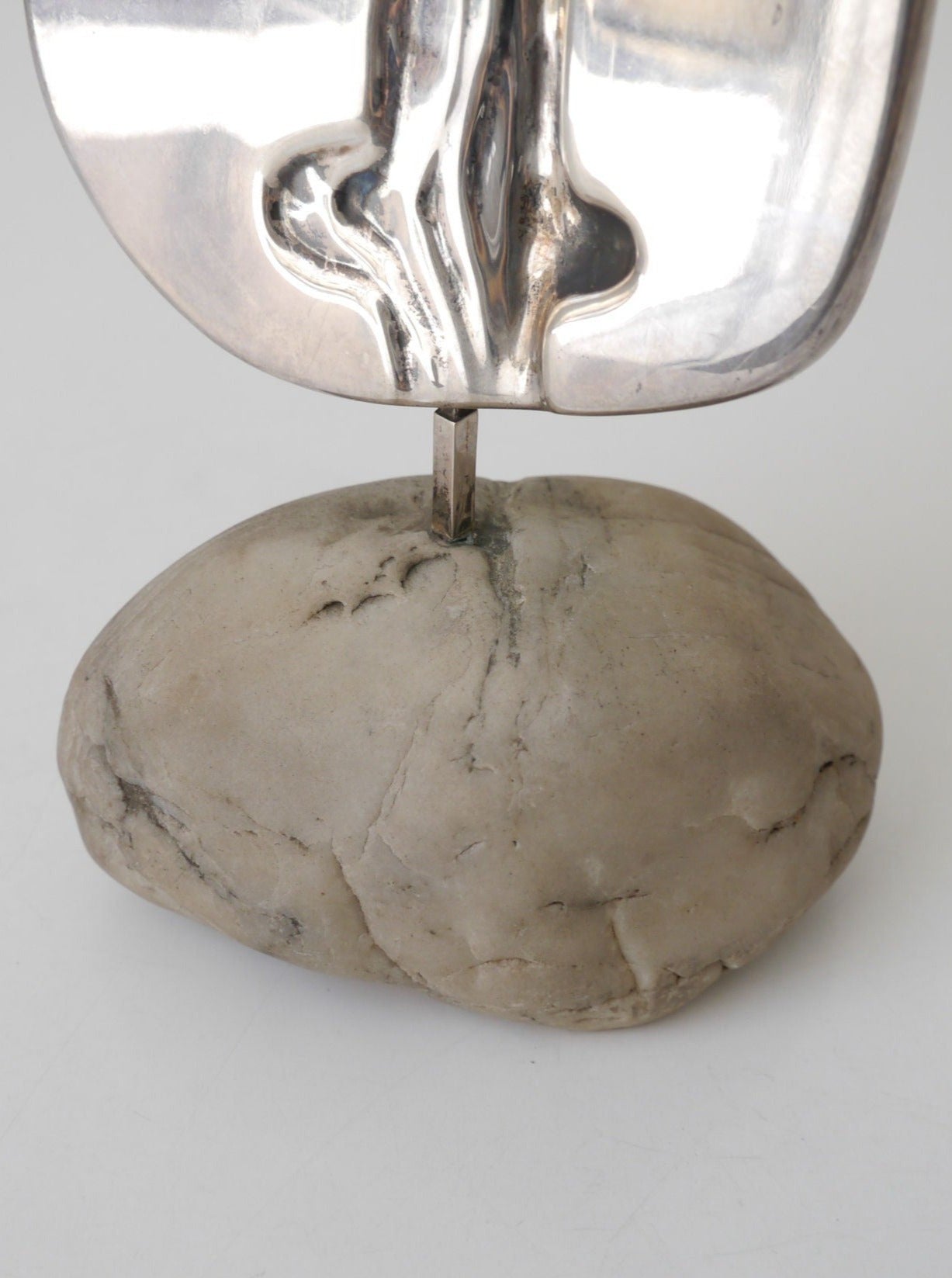 A sculptural piece featuring a shiny De Vecchi Milano Silver Sculpture mounted on a rough, gray stone with visible cracks. The metal has a reflective, contoured surface from Collection Apart.