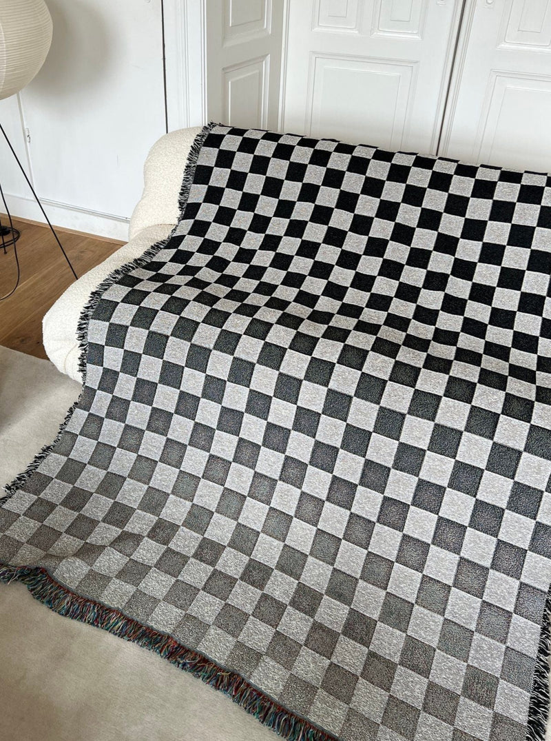 A black and white checkered Goto Jacquard Throw from 2222Studio is draped over a white couch in a room with decorative white paneling on the wall.