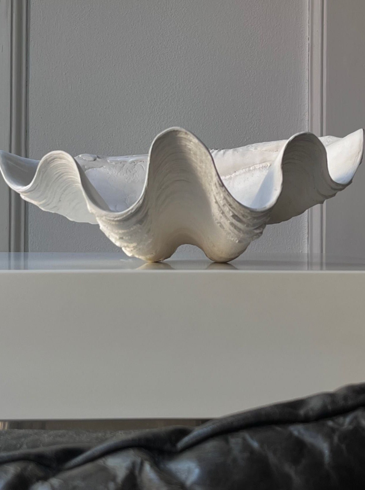 A wavy, sculptural white Médecine Clam Shell Bowl with an undulating rim, placed on a shelf against a soft grey background, highlighted by natural light.