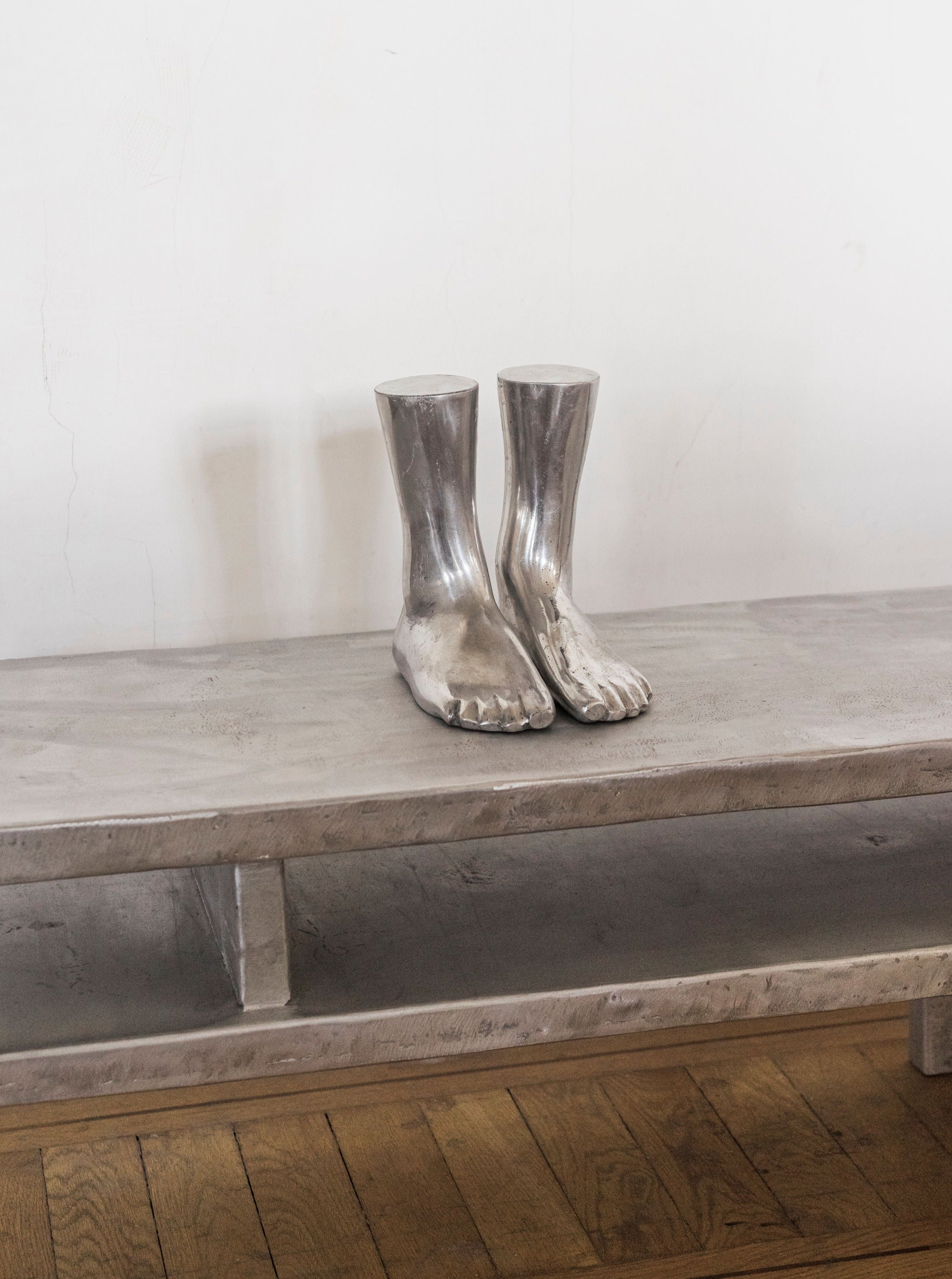 A sculpture of Rooms Studio's Aluminum Feet Sculpture fused with human feet displayed on a wooden bench against a white wall with visible cracks.