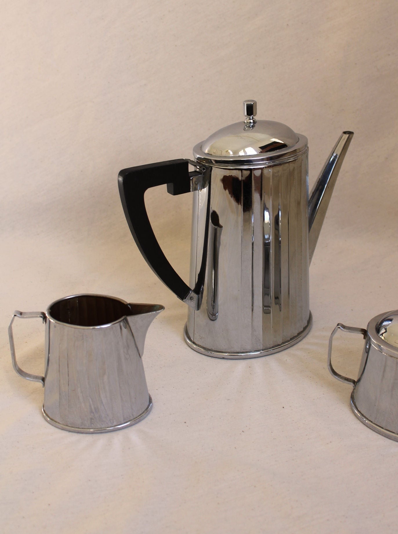 A shiny stainless steel Art Deco Coffee Set by Maison Collectible with a black handle, displaying timeless elegance, accompanied by two smaller matching creamers on a beige surface.