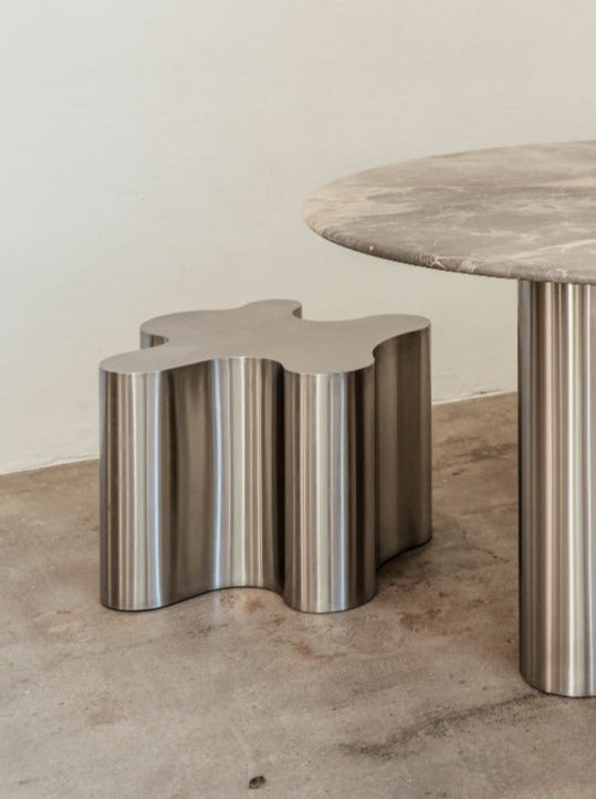A modern Silver Root - Low stool with a puzzle piece-shaped seat, made of shiny brushed metal, stands next to a round, column-like concrete table with a sculptural steel base in a minimalist setting by Caia Leifsdotter.