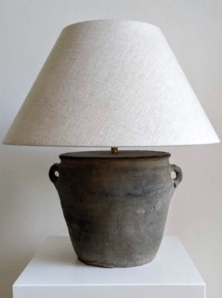 An antique Médecine earthenware lamp attached to an old, rustic clay pot with handles, set on a white pedestal against a plain background. The pot lends a unique base to the otherwise traditional lamp.