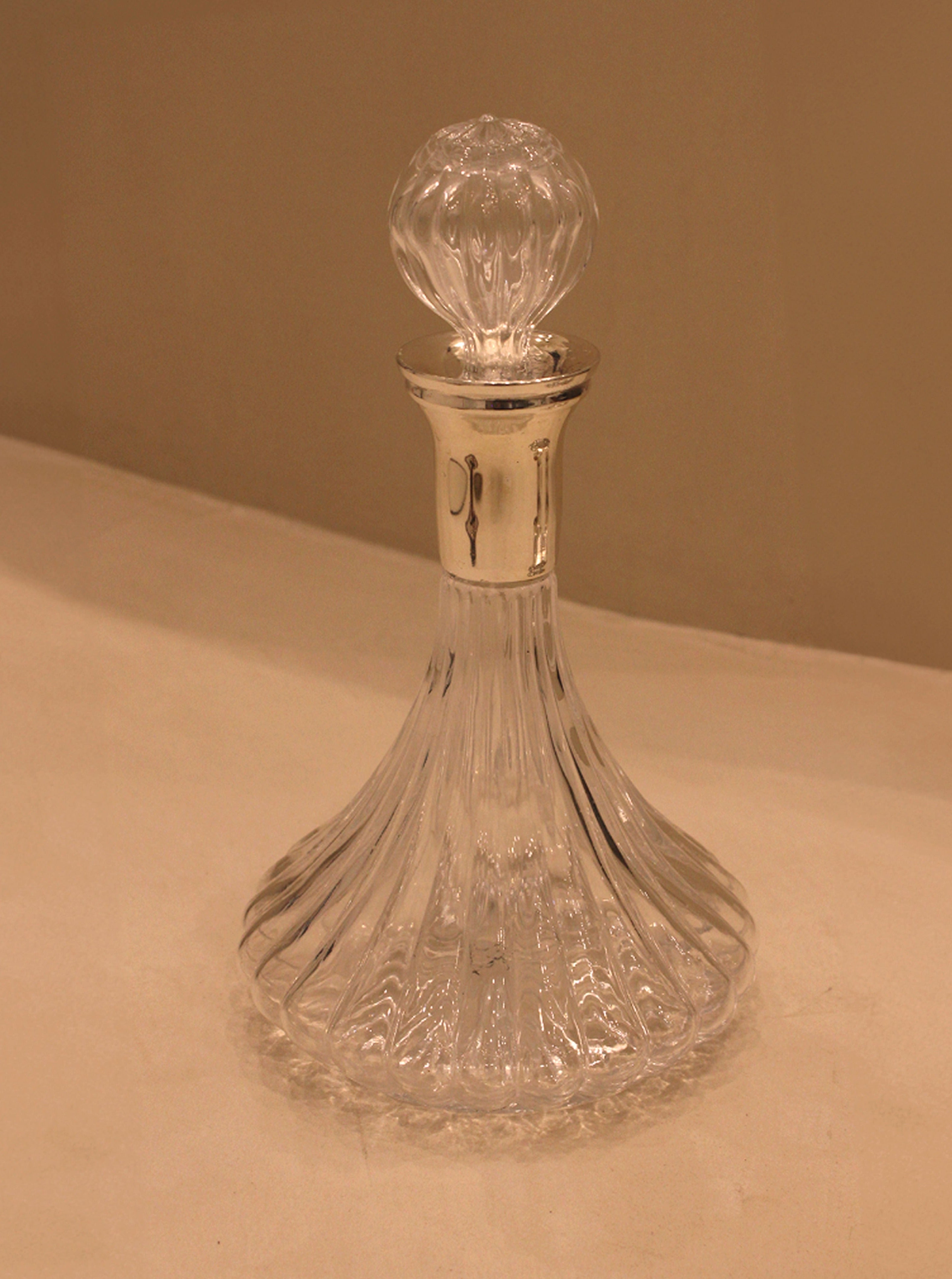 A Maison Collectible clear glass carafe with an elegant fluted base and a rounded stopper, displayed on a beige surface under warm lighting.