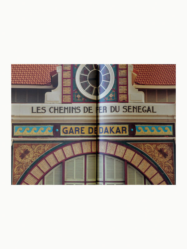 Decorative facade of the Dakar railway station labeled "Les Chemins de Fer du Senegal, Gare de Dakar" with intricate designs and terracotta roof tiles; center obscured by a Maison Plage: Julian Schnabel: CVJ: Nicknames of Maitre D's & Other Excerpts from Life.