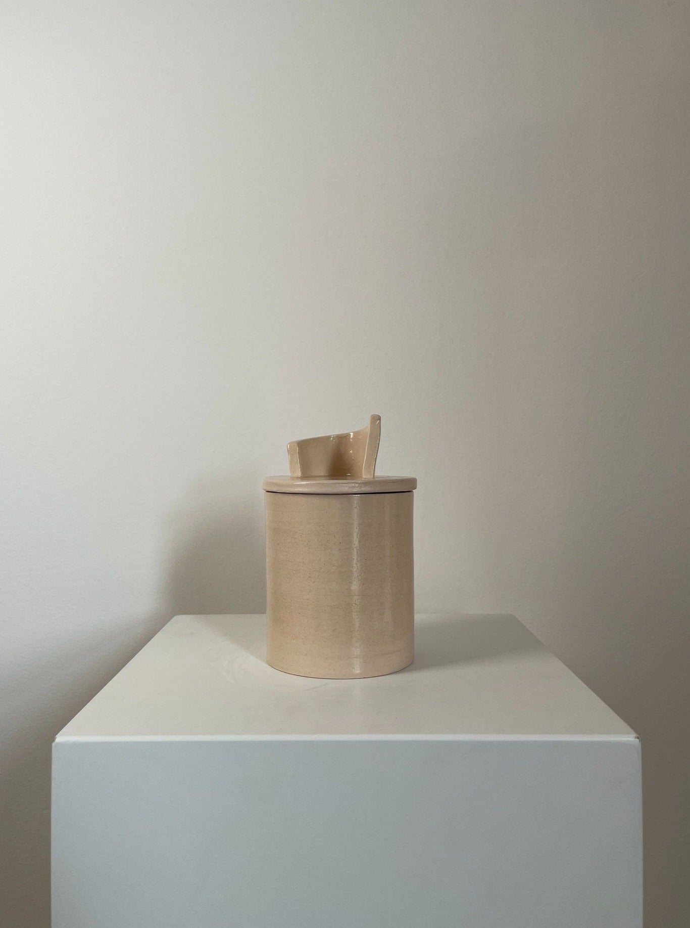 A wooden container with a lid, placed on a white pedestal against a plain light-colored wall. The lid is slightly ajar with a 2222Studio Sculptural Scented Candle No. 11 peeking out.