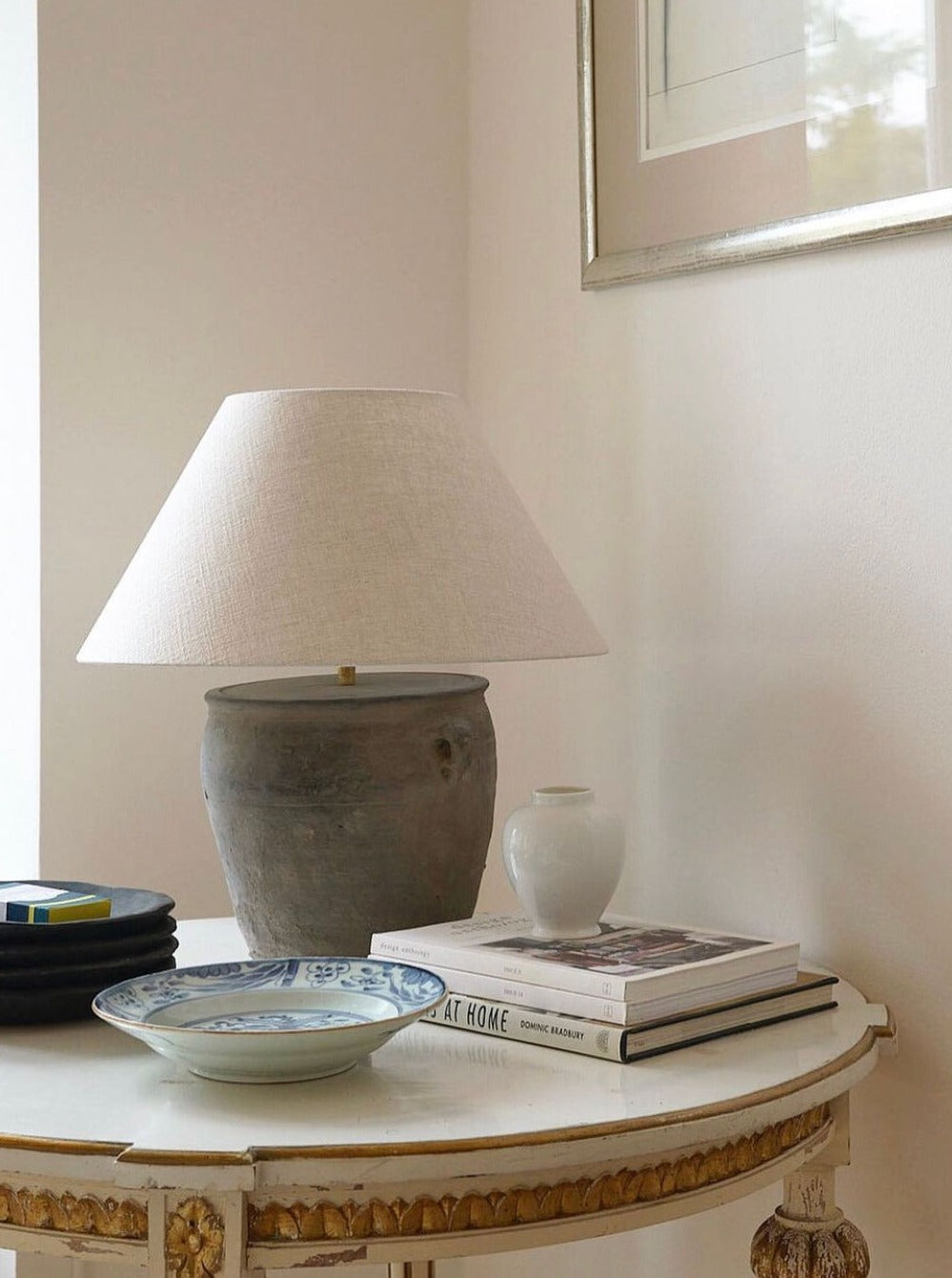 A cozy corner featuring a round table adorned with a Médecine antique earthenware lamp with a white shade, a stack of books, a small white vase, and a decorative blue and white plate.