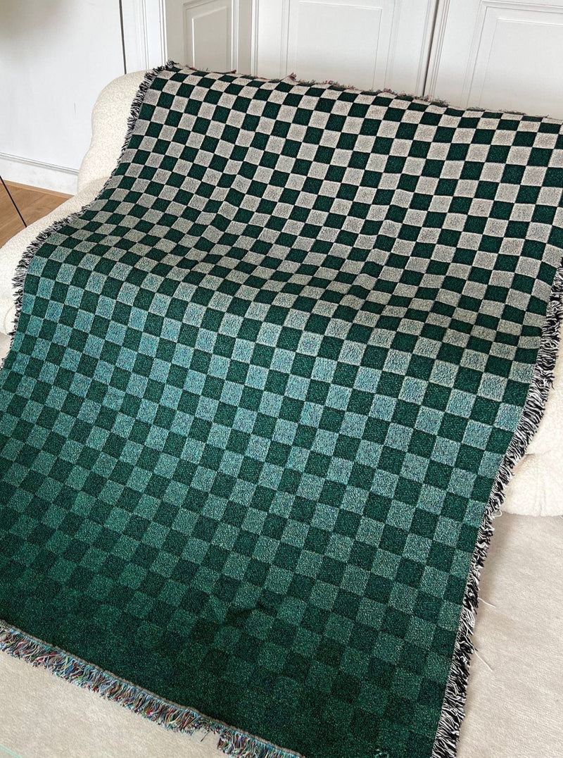 A dark and light green checkered Tori Jacquard Throw draped over a white couch, displaying a cozy and textured look with fringed edges from 2222Studio.