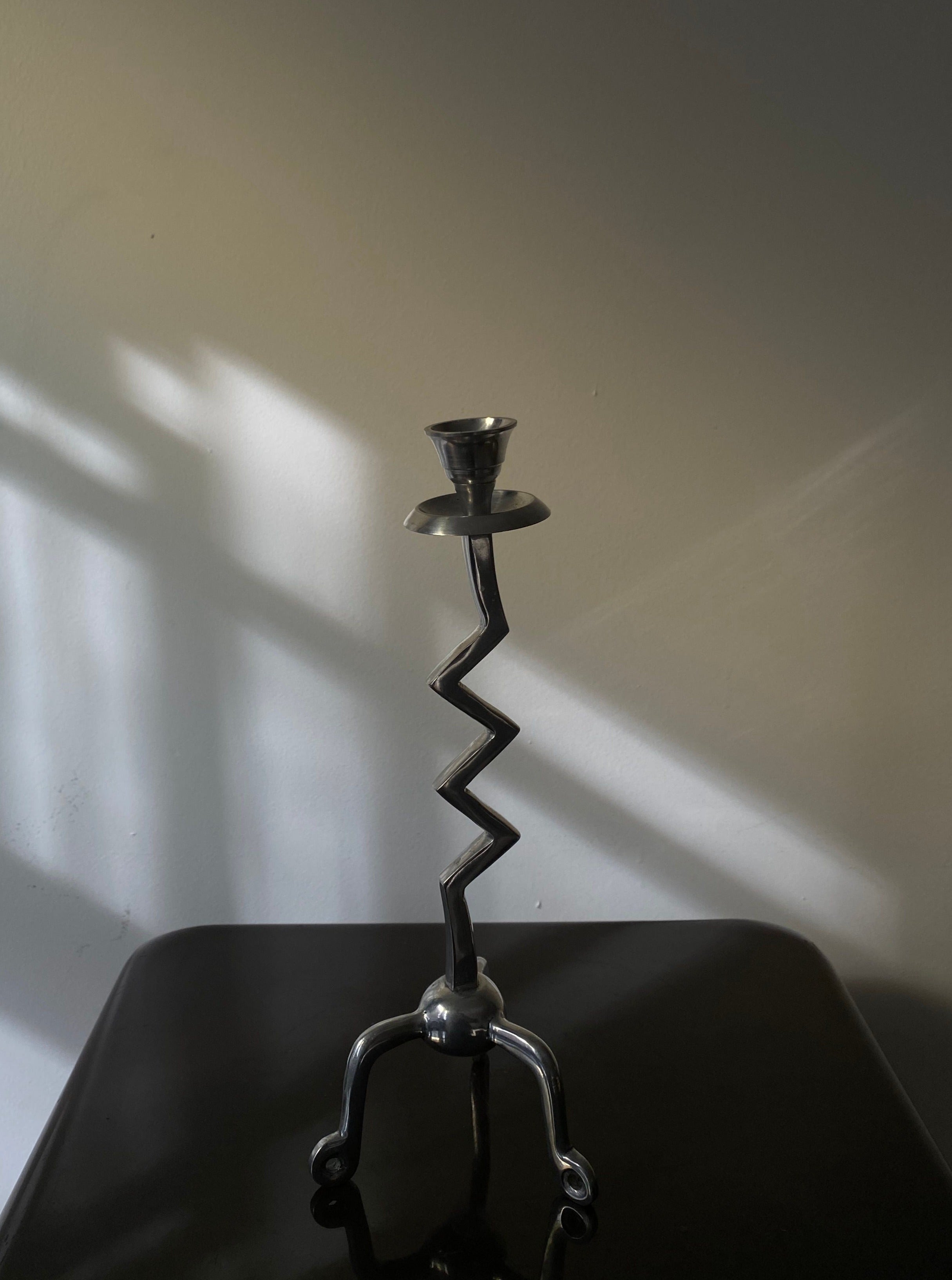 A Les Objets Amsterdam vintage zigzag candle holder with a tripod base, positioned on a dark surface against a light wall with shadow patterns.